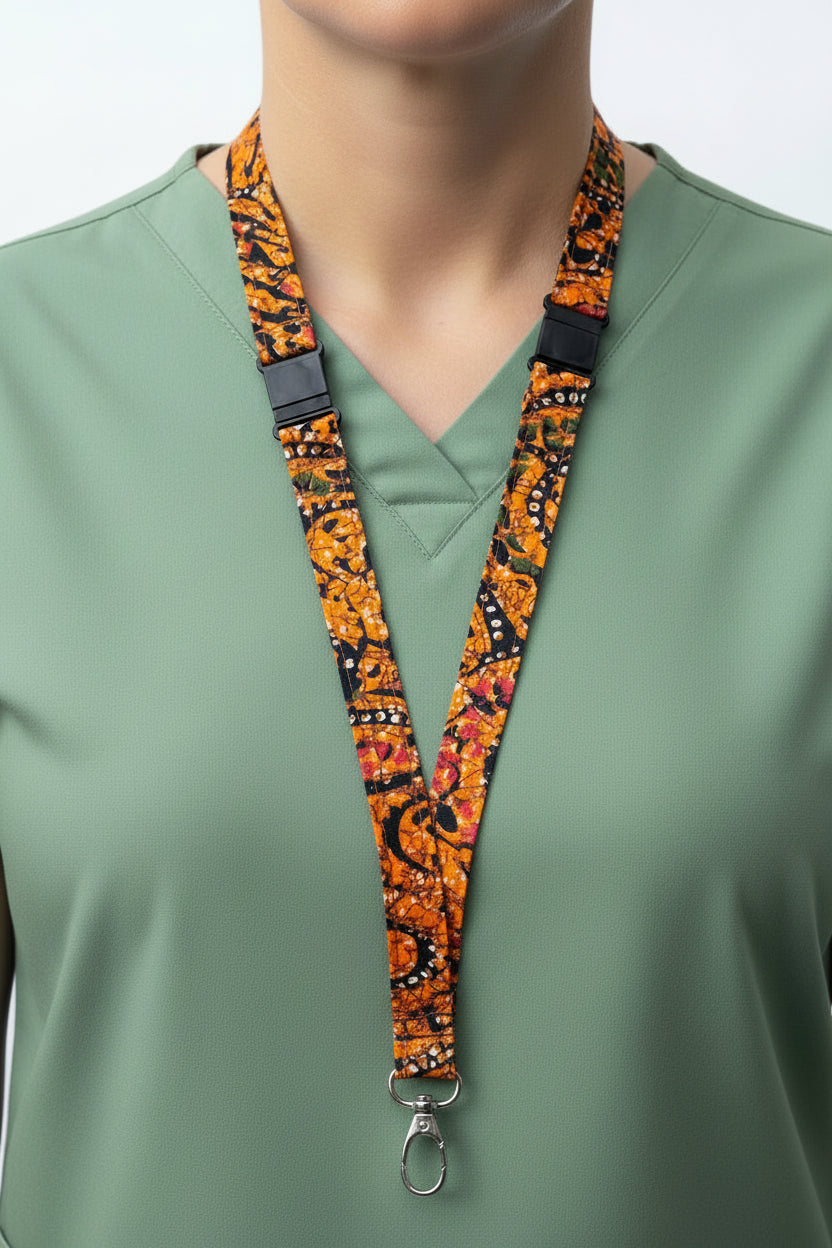 A close-up of a patterned lanyard worn around a light gray turtleneck sweater. The lanyard has an orange, black, and red paisley-like print with a black plastic buckle and a silver metal clip at the bottom.